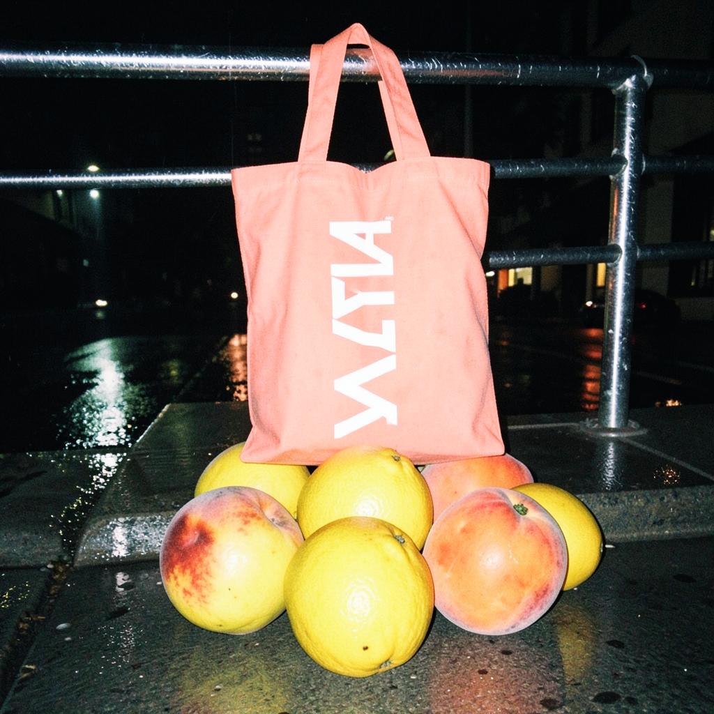Coral ZYLA tote bag hanging on a railing above a pile of giant peaches and yuzu